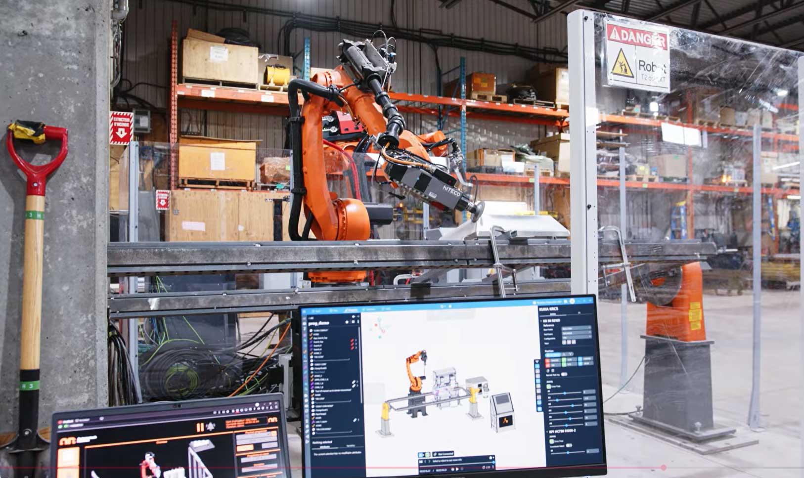 Industrial robotic arm welding inside automated cell with programming interface visible on nearby screen.